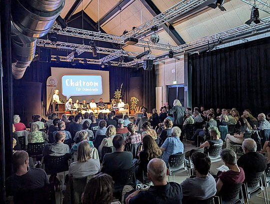 Gesamtansicht der Halle mit Publikum und Bühne mit Kandidat*innen aufgenommen von der hinteren Reihe