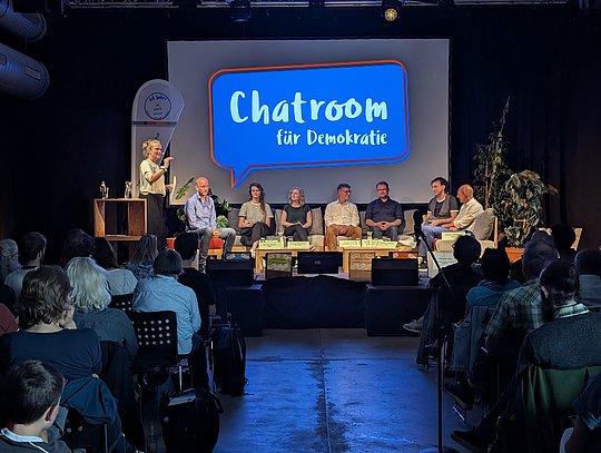 Bühnenansicht mit allen Kandidate*innen und der Diskussionsleiterin aus der Sicht des Publikums